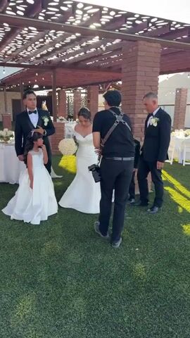 The video shows a wedding photoshoot outdoors. A photographer, wearing a black shirt and pants with a harness holding two cameras, is standing on a grassy lawn. To the left, a young girl in a white formal dress and a man in a tuxedo are visible near a round table. To the right, two young boys in tuxedos and a man in a tuxedo are standing. A bride in a white wedding dress is in the background, facing away from the camera. The setting appears to be a venue with a covered patio and tables set up for an event. Sunlight is casting shadows on the lawn. The camera then shifts to a close-up of a hand holding a camera lens.