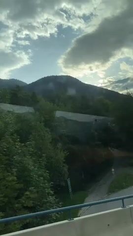 The video shows a view from a vehicle, possibly a train or bus, moving alongside a forested hillside under a cloudy sky. The hillside is covered in lush green trees and bushes, with a mountain peak visible in the distance. A road or path winds through the lower part of the scene, and there are some signs and what appears to be a construction area with white coverings. The sun is partially visible through the clouds, casting light on the landscape. The camera is panning from left to right, capturing the changing scenery.