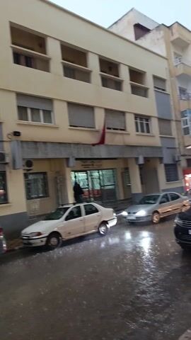 The video depicts a rainy street scene in what appears to be a city. Several multistory buildings line the street, with parked cars along the curbs. The weather is overcast. Several cars are seen driving down the street, with water splashing. A person is seen entering a building in the beginning of the video.