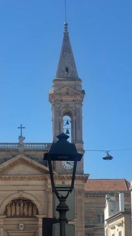 The video shows a view of a church tower and its facade on a clear blue sky. A black lamppost is in the foreground, partially obscuring the view of the church. The church tower is made of stone and has a steeple. Bells are visible inside an arched opening in the tower. A clock face is also visible on the tower. To the left of the tower, a cross is mounted on a balustrade above the church facade. The facade is adorned with sculptures. To the right, part of another building with a balcony is visible. The video pans slightly and zooms in on the church tower. The timestamp indicates the video starts at 00:00 and ends at 00:04.