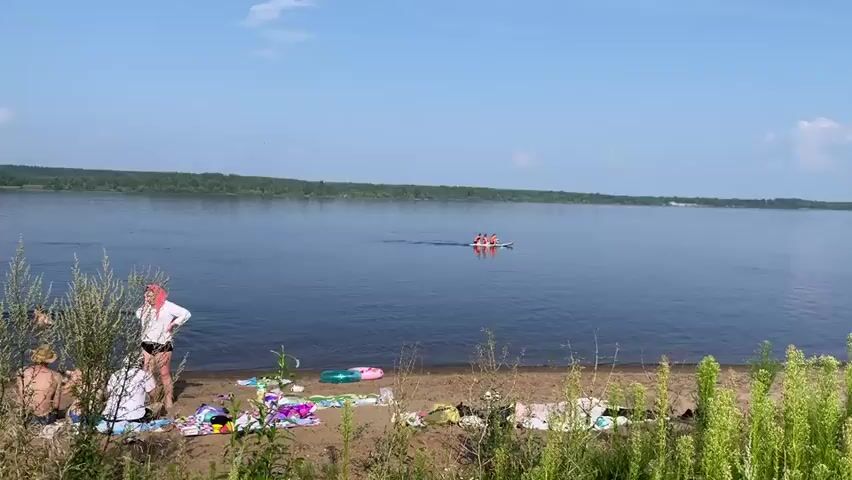 A group of people are gathered on a sandy beach by a wide river on a sunny day. Some individuals are in the water, while others are sitting on the sand near towels and beach gear. In the background, several people are on a paddleboard in the river. The river is calm and wide, with a tree line visible on the opposite bank. The sky is blue with scattered clouds.