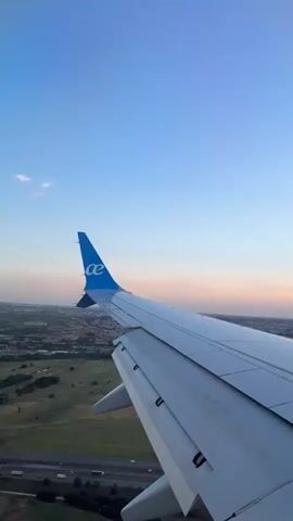 The video shows an airplane wing in flight, seen from inside the plane. The wing is white with a blue tail fin displaying a white logo. Below, a landscape of green fields and buildings stretches out, intersected by a highway with moving traffic. The sky transitions from blue to a light pinkish hue near the horizon. An electrical tower can be seen in the lower frame.