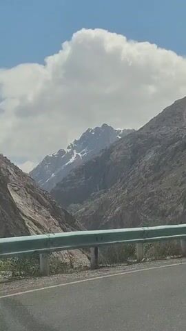 The video shows a car driving on a paved road next to a mountain range. A guard rail is visible on the side of the road. Some of the mountains have snow on them. The sky is blue with white clouds. There is music playing in the background.