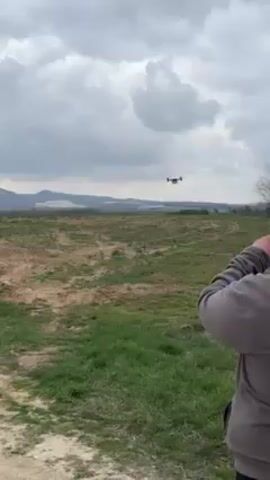A person stands at the edge of a grassy field, watching a drone fly towards them. The drone is white with black rotors. The field is mostly brown, with patches of green grass. In the distance, hills and mountains are visible under a cloudy sky.
