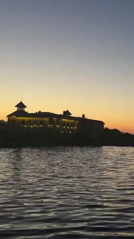 The video pans across a body of water reflecting the warm hues of a sunset. In the distance, a silhouetted building with illuminated windows sits on the horizon, surrounded by dark trees. The sky transitions from a deep blue at the top to a vibrant orange and yellow band just above the horizon. The water is calm, with gentle ripples disturbing its surface, and a faint line of what appears to be a barrier or a distant shore with posts is visible across the water. The overall scene is serene and atmospheric, capturing the beauty of twilight.