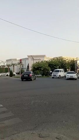 The video displays a street scene in Termez, Uzbekistan. The time of day appears to be late afternoon or early evening, as suggested by the soft, warm light and the clear sky. The weather seems pleasant, with no visible signs of rain or strong winds.

The main focus is a busy intersection in front of a large, modern building with a sign that reads "SERENADA". The architecture of the building is contemporary, with a white facade, large windows, and decorative elements. Palm trees and lush green trees are planted around the building, adding to the aesthetic.

Several vehicles are present in the scene, including cars and a small white truck. Traffic lights are visible, indicating the presence of an active intersection. A pedestrian crossing is also in view, with a white car approaching it. In the background, a billboard advertises a product, displaying text that appears to be in Cyrillic or a similar script, along with a price of "109 990".

The overall atmosphere of the video is one of everyday urban life. There are no discernible emotions conveyed as the focus is on the movement of traffic and the general environment. The scene suggests a commercial or entertainment district, given the size of the "SERENADA" building and the billboard. The presence of multiple cars and the structured intersection point towards a developed urban area.