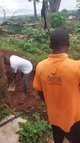 The video shows a group of people gathered outdoors on a dirt and grassy slope. The foreground features the back of a person wearing an orange polo shirt with a logo that reads "Astral Water Pump". In the midground, several other individuals are visible. One person in a blue shirt is speaking with their hands outstretched, while others in the background are standing and observing. The environment appears to be somewhat overgrown with vegetation and trees, and there is a brown wall or building visible in the background. The overall impression is of a casual outdoor gathering or discussion.