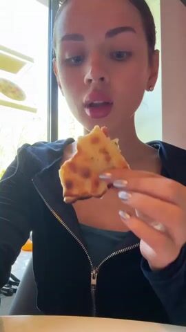 A young woman is eating a piece of cheese bread indoors. The woman is light-skinned with brown hair and eyes, and she has on silver nail polish. She wears a blue top with a partially-zipped dark blue velvet jacket. The piece of bread appears to have cheese melted on top, and the woman takes a large bite. A thin string of cheese stretches between the bread and her mouth. The environment is an indoor dining area, and there is a window visible with natural light.