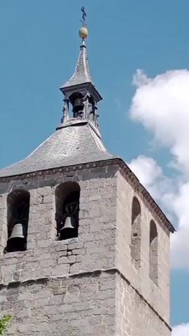 The video shows the top of a stone bell tower with bells visible in arched openings. The tower is topped with a steeple and a cross. The sky is blue with some white clouds. The camera pans slightly upwards.