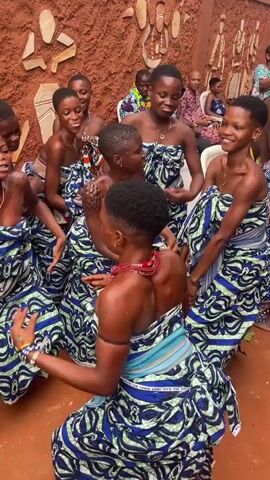 A group of young women, dressed in matching blue and green patterned wraps, are dancing in a circle. They appear to be in an outdoor setting with a textured, reddish-brown wall behind them adorned with carved or painted figures. Some of the women are looking at each other and smiling, suggesting a celebratory or cultural event. Their skin is glistening, possibly from sweat or water, and they are adorned with jewelry, including necklaces and armbands. The lighting suggests it is daytime.