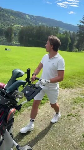A young man in a white polo shirt and shorts pushes a golf cart across a green lawn. He is looking to his right. The golf cart is filled with clubs. In the background is a lake and a wooded mountain. There are some markers on the lawn in the distance. The sky is blue with some clouds.
