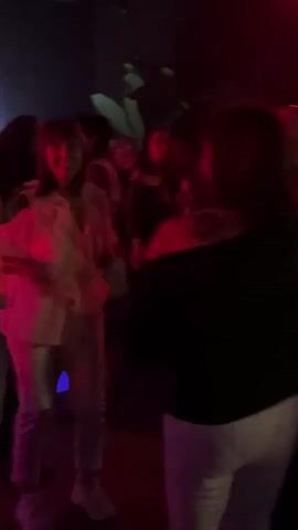 The video shows a group of people dancing in what appears to be a club or party setting. The lighting is dynamic, shifting between blue and red, adding to the energetic atmosphere. Several individuals are visible, engaging in dance moves. In the background, a DJ is positioned behind a console, presumably responsible for the music that drives the scene.