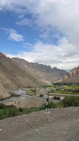 A video shows a scenic mountain valley with a river, a road, and a small village. The sky is blue with scattered clouds. In the foreground, there are concrete barriers with red and white markings, and traffic lights with red and yellow lenses. A pole with colorful tape is also visible. Several vehicles, including cars and a truck, are on the road. The valley is surrounded by barren, rocky mountains. The village appears to have several buildings and fields, some of which are yellow. The camera pans across the landscape, offering different views of the valley and the surrounding mountains.