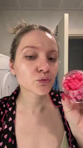 A woman with blonde hair in a bun and wearing a black pajama top with pink hearts is in a bathroom. She has a nose ring and is applying a product from a clear jar filled with small pink capsules to her face. She uses a small spatula to scoop out the product and applies it to her cheek. A washing machine is visible in the background.