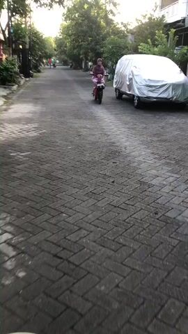 The video begins with a low-angle shot of a brick-paved road. In the distance, a woman wearing a hijab rides a motorcycle. To the right of the road, a car is parked, covered by a silver tarp. The road is lined with trees and vegetation, suggesting an outdoor setting. The lighting indicates it may be early morning or late afternoon. The camera is in motion, tracking the motorcycle rider.