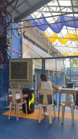 The video shows an indoor sports facility. The ceiling is covered with blue and yellow fabric draped in an undulating fashion. Metal beams and a corrugated metal roof are visible. A large white industrial fan or air cooler unit with the brand name "IWATA" is positioned to the left of the frame, next to a black speaker with a green light. Three plastic chairs are arranged in front of a metal table, and a woman wearing a white top and denim shorts is sitting on the middle chair, facing away from the camera. She has her left arm resting on the back of the chair.

In the background, a blue court marked with orange and white lines is visible through a metal fence. Two people are playing a sport on the court, possibly pickleball, indicated by the presence of paddles. One person is wearing a light blue shirt and black shorts, while the other is wearing a green shirt and black shorts. Another individual in a black shirt is standing near the court. Further back, a large banner with text and graphics is displayed on the wall. Several people are sitting on the floor to the right of the frame, watching the game. They are wearing casual clothing. A white ladder is leaning against the right side of the fence. The flooring of the facility appears to be a blue padded mat with orange lines.