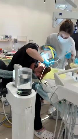 A man lies reclined in a dental chair, wearing black sunglasses and a blue paper surgical cap. A dentist, wearing a black shirt, blue gloves, a surgical mask, and a clear face shield with a yellow band, leans over the patient, working inside his mouth with dental instruments. A dental assistant, wearing a white shirt, blue gloves, and a surgical mask, stands to the patient's right, holding a dental tool. The dental office appears well-lit and sterile, with various medical equipment visible in the background, including a dental unit with a water reservoir and a light fixture above the patient's head.