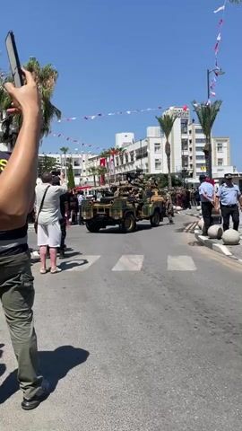 A military parade is taking place on a sunny day in a town. Several military vehicles, including jeeps and a truck, are moving down a street lined with palm trees and buildings. Soldiers in camouflage uniforms are visible in and on the vehicles, some carrying weapons. Spectators, including people taking photos, are standing on the sidewalks, and police officers are also present. Red and white flags are strung across the street. The buildings appear to be hotels. The overall atmosphere seems festive and orderly.