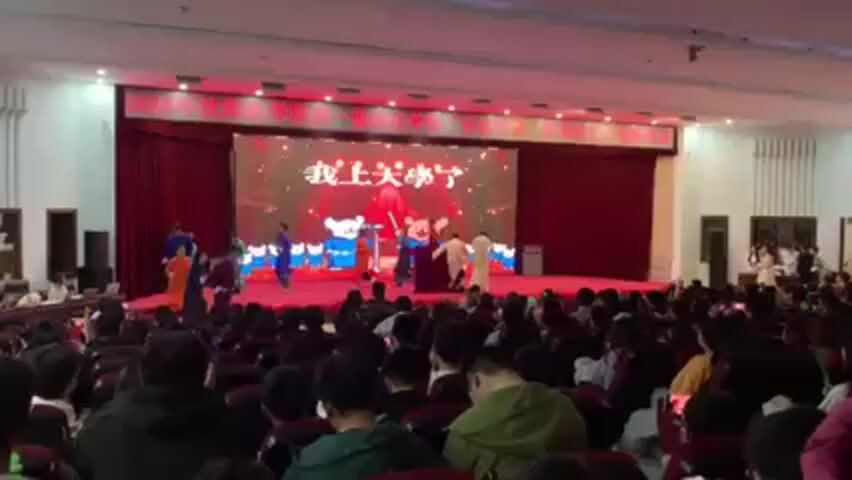A stage show appears to be occurring in an auditorium filled with audience members. The stage features a backdrop with Chinese characters and a cartoon bear design. Performers in various colorful costumes are dancing on stage. Many audience members are holding up their phones to record the performance. The auditorium has red seating, bright lighting, and simple decorative elements.