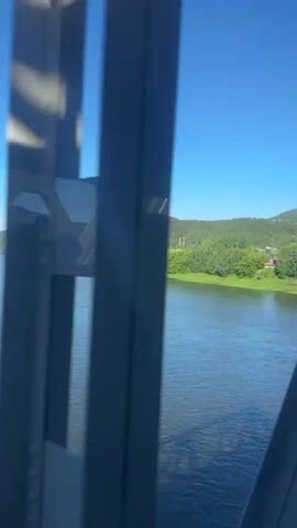 This video captures a scenic view from a bridge overlooking a wide, calm river in Ust-Mana, Russia. The time of day is clearly midday, indicated by the bright, clear blue sky and the strong sunlight casting shadows. The weather is fair and warm, with no visible clouds.

The camera is positioned on a metal bridge, with its dark, riveted structure dominating the foreground. A reflection of what appears to be a person taking the video can be seen in a small, rectangular window on the bridge's structure. The bridge's construction is industrial, with thick metal beams and numerous rivets.

The river itself is a deep blue, with gentle ripples on its surface, suggesting a slow current. Along the far bank of the river, a lush green landscape unfolds. Dense forests of deciduous trees cover rolling hills, interspersed with some open, grassy areas. The greenery is vibrant, indicating a lush growing season, likely late spring or summer.

Small clusters of buildings, likely a village or settlement, are visible nestled amongst the trees along the riverbank. These buildings appear to be residential, with some having red roofs. The overall impression is one of a peaceful, rural setting.

The video pans slowly, offering a panoramic view of the river and the landscape. The activity taking place is simply the traversal of the bridge, allowing for the observation of the surrounding natural beauty. There is a sense of tranquility and appreciation for the natural environment. No text is visible in the video.