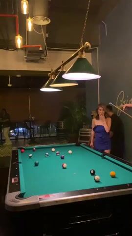 The video shows a group of people playing pool in a bar. The pool table is green, and the balls are scattered across it. There is a woman in a blue dress standing next to the table, and a man behind her appears to be giving her advice. The bar has a casual and relaxed atmosphere. There are barrels near a potted plant on the right side of the pool table. The hanging light fixture over the table has three green lampshades. A man in a black t-shirt stands by a high table, next to a window.