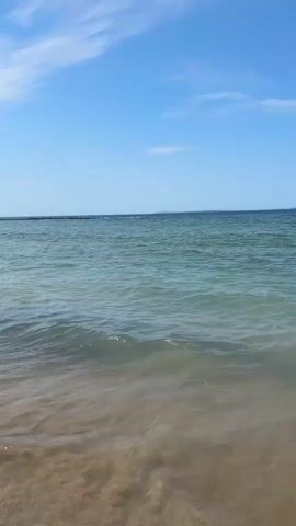 This is a vertical video of a serene beach scene. The camera is positioned at a low angle, showing the gentle waves lapping onto a sandy shore. The water transitions from a clear, shallow sandy bottom near the viewer to a deeper, turquoise green hue further out. In the distance, a faint, dark landmass is visible on the horizon. The sky above is a bright, clear blue, dotted with a few wispy white clouds. Towards the end of the clip, a person is briefly visible swimming in the water on the right side of the frame. The overall atmosphere is peaceful and tranquil.