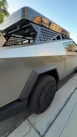 The video shows a side view of a Tesla Cybertruck parked on a street next to a curb. The truck is silver with black accents and has a rooftop rack with a tent or cargo box. The sun is shining, casting reflections on the truck's surface. The background shows houses and palm trees. The camera pans slightly to the right.
