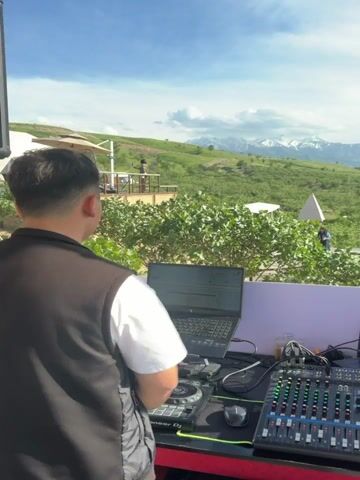 A DJ is setting up at an outdoor event. The DJ, wearing a vest over a white shirt, has his back to the camera and is working on a laptop and mixing board. In the background, there is a wooden deck with people dressed in casual clothing, seemingly enjoying the view and taking pictures. The environment is a lush, green landscape with rolling hills, and in the distance, there are snow-capped mountains under a blue sky with scattered clouds. There is a white tent and a white pyramid-shaped structure on the landscape.