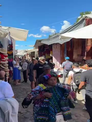 The video shows a bustling outdoor market on a sunny day. People of various ages and ethnicities stroll through the narrow passageway between stalls covered by white canopies. Many of the people appear to be tourists. The stalls display colorful textiles, hats, and various crafts.  Some individuals carry bags or backpacks.  One woman in the foreground wears traditional indigenous clothing and carries a folded textile. The ground appears to be paved with cobblestones. The sky is blue with scattered clouds.