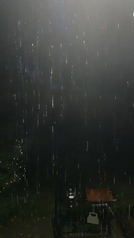 The video shows a dark, outdoor scene with rain falling. The rain is illuminated by a bright light source at the top of the frame, creating streaks of light as the drops descend. In the background, a Christmas tree decorated with lights is visible on the left. A grill with a wooden surface is positioned on the right side of the frame. The ground appears wet, suggesting accumulated rainwater. The overall atmosphere is dark and moody, with the rain and lighting creating a dramatic effect.