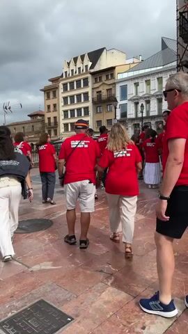 A group of people wearing matching red t-shirts walk across a plaza paved with reddish-brown tiles. The t-shirts appear to have writing on the back. In the background, there are several multi-story buildings with a variety of architectural styles. To the left, a tall, ornate cathedral with a spire dominates the skyline. The sky is overcast. A person in sunglasses and a dark shirt walks into the frame from the right.