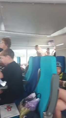 The video shows the interior of a passenger vessel. Several people are seated in blue and turquoise seats. A young boy in a blue hat is looking towards the camera. In the background, a woman with blonde hair is seated. Further back, a man in a white shirt is standing near a window, and a woman with dark hair is also standing. The lighting appears to be natural light coming from the windows. The overall atmosphere seems casual and relaxed.
