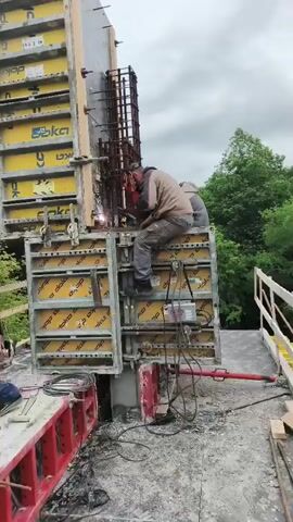 Two construction workers are seen at a construction site. One worker, wearing a tan jacket and grey pants, is positioned on top of a large yellow metal formwork, welding metal rebar. He is wearing a welding mask. Another worker, wearing a grey jacket and blue jeans, stands to the right of the formwork, his arm resting on it. He is wearing a red hard hat. The formwork, which is tall and rectangular, is made of yellow panels with black markings. Wires and a welding machine are visible at the base of the formwork. The background shows green trees and an overcast sky. A red metal structure is partially visible in the foreground.
