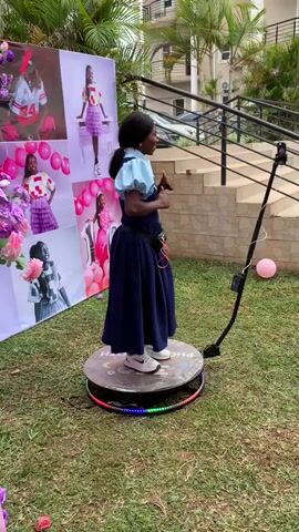 A young Black woman stands on a spinning platform in a grassy yard, facing a backdrop with multiple photos of her. The platform is circular and has a ring of colorful LED lights around its base. A camera on an adjustable arm is positioned in front of her. She is wearing a dark blue dress with a light blue collar and cuffs, white sneakers, and a fanny pack. Her hair is pulled back. She is smiling and gesturing with her hands. The backdrop features images of the woman in various poses and outfits, interspersed with pink balloons and floral decorations. In the background, there is a building with stairs and palm trees. A pink balloon is visible on the grass to the right of the platform. The video captures the woman posing and interacting with the camera as the platform rotates.