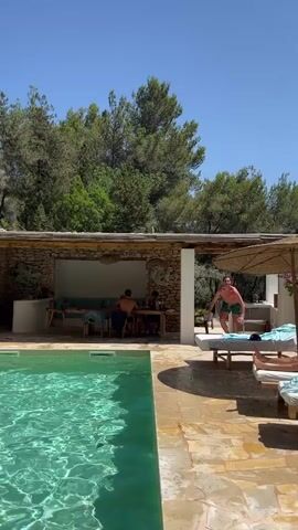 A man in green swim trunks jumps from a covered patio area with a stone wall into a bright blue swimming pool. He is in mid-air, performing a flip with his arms and legs extended. The pool is surrounded by a stone patio and lounge chairs are visible in the background. Trees and a clear blue sky form the backdrop. Several people are seated at a table under the covered patio.