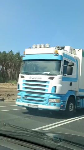 The video captures a roadside view of a construction site next to a pine forest under a clear blue sky, suggesting a sunny day. The scene is divided by a highway with white lines marking lanes.

The primary subjects are construction vehicles. A large dump truck with a yellow and white cab drives on the highway, while another dump truck with a darker cab is seen on the construction site. Two excavators, one orange and one yellow, are actively working on the dirt and sand mounds of the construction area. A striped red and white construction marker is also visible near the highway.

The activity is construction, likely roadwork or land development, given the excavators and dump trucks. The vehicles are moving and operating, indicating work is in progress. The presence of the forest and the clear sky sets the scene in a rural or semi-rural environment.

There is some visible text on the side of one of the dump trucks, appearing to be a company name or logo, though it is partially obscured and difficult to read clearly. The overall mood is functional and industrial, reflecting the ongoing construction work. The camera appears to be mounted inside a vehicle, looking out, as evidenced by the dashboard in the foreground.