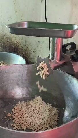 A close-up, eye-level shot shows a commercial meat grinder in operation. The grinder, made of stainless steel, is positioned over a large, silver metal bowl. Ground meat, light beige in color and appearing somewhat moist, is being extruded from the front of the grinder in a continuous stream, falling into the bowl and accumulating. The meat has a rough, granular texture. The grinder has a feeding tray at the top and a handle or knob on the side. The background is out of focus, but appears to be a wall with some staining and a black electrical cord hanging. The overall lighting suggests an indoor, possibly industrial or commercial kitchen setting.