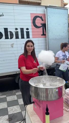 The video shows two women standing outside. The woman in the foreground is operating a cotton candy machine. The machine is pink and has a large stainless steel bowl on top. She is using a wooden stick to collect the cotton candy, creating a large, fluffy white cloud. She is smiling and appears to be enjoying the process. She is wearing a red t-shirt and grey jeans.  Behind her is a large white truck with black lettering on the side. Another woman is standing behind the cotton candy machine, pouring liquid from a white container. She is wearing a white t-shirt, grey trousers and has her hair in a ponytail.  The ground is covered in black and white checkered tiles. The environment appears to be an outdoor event or festival.