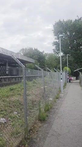 The video shows a chain-link fence in the foreground with overgrown grass and weeds. Beyond the fence, there is a train platform with a covered waiting area. The platform appears to be old and somewhat dilapidated. The camera then pans to the right, revealing a set of outdoor stairs with yellow-striped edges and metal railings. The stairs lead upwards towards an overhead structure, possibly a walkway or bridge. The sky is overcast, and trees are visible in the background.