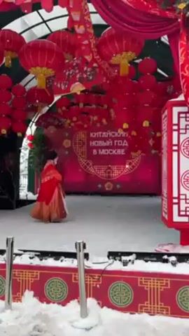 The scene captures an outdoor celebration of the Chinese New Year in Moscow, Russia, during winter. A person dressed in a red and golden traditional Chinese costume stands on a snow-covered stage. The stage is decorated with numerous red lanterns hanging from above, and the backdrop features a sign in Russian that reads "Китайский Новый Год в Москве," meaning "Chinese New Year in Moscow". A decorative red and white lantern stands to the right of the stage, and snow falls lightly, adding to the winter atmosphere.