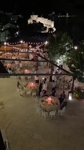 An aerial shot shows an outdoor evening reception with multiple round tables set with white tablecloths and chairs. Many guests in light-colored clothing are mingling and sitting at the tables, some of which are lit by candles. String lights are draped overhead, illuminating the area. In the background, a fortified structure sits on a hill overlooking a body of water at night.
