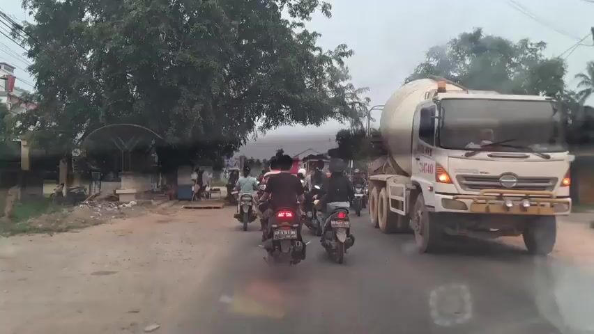 The video shows a view from a vehicle moving on a road. The road is paved and appears to be in a somewhat urban or semi-urban area. On the right side of the road, a large concrete mixer truck is prominently visible. It is a white truck with a rotating drum, and it has "MITRA MIX" written on its side. The truck has several wheels and appears to be moving. In front of the truck and also on the road, there are several motorcycles. These motorcycles are carrying riders, some of whom are wearing helmets. The motorcycles are moving in the same direction as the vehicle from which the video is being recorded. On the left side of the road, there is a large tree with dense foliage. Below the tree, there is a roadside structure that appears to be a shelter or a small stall with some people standing around it. Further down the road, there are buildings visible, including a multi-story building with a light-colored facade. The sky is overcast, suggesting it might be dawn, dusk, or a cloudy day. The overall scene depicts traffic on a road with a mix of vehicles and pedestrians in a roadside environment.