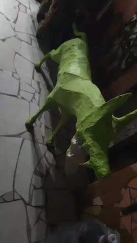 A green paper mache animal sculpture resembling a dog or wolf is attached to a wall covered in a mosaic of light and dark triangular tiles. The sculpture is positioned vertically, with its head facing downwards and its body angled away from the wall. Its mouth is open, revealing white teeth and a pink interior. Two front legs are bent and appear to be gripping the wall, while the hind legs are also bent and angled downwards. The sculpture's body is elongated and textured, with visible seams from the paper mache. To the lower right of the sculpture, a large clear plastic water jug with a red and white cap is partially visible, resting on the floor. The background includes elements of what appears to be a rough, textured wall or a decorative element with dark, spiky textures.