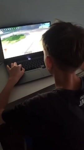 A person is playing a video game on a laptop. The scene is from behind the person, showing their head and shoulders as they are focused on the screen. The laptop is open on a desk, and the person's left hand is resting on the keyboard. The game appears to be a 3D world with a cartoony art style, featuring green platforms and a light-colored background. The person is wearing a dark t-shirt with some text on the back, but it is not clearly legible. The lighting suggests it could be daytime or the room is well-lit. The setting seems to be indoors, likely a home environment. There is no discernible emotion conveyed, other than concentration on the game. The timestamp indicates the video is from an unknown time.
