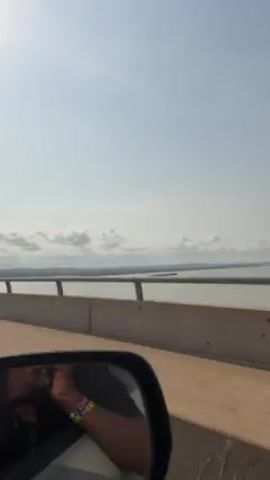 The video shows a view from a moving vehicle, likely a car, crossing a bridge. The focus is on the landscape outside the car window. A wide body of water is visible, with land in the distance. The sky is overcast with puffy clouds. The bridge has a gray concrete barrier and a metal railing. In the car's side mirror, a person's arm with a multi-colored bracelet can be seen. The car is moving along the bridge.
