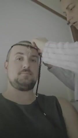 A man with short brown hair and a beard is sitting while someone is braiding his hair. A decorative accessory is woven into the braid on his forehead. A person, mostly out of frame, is using their hand to cover the man's eyes, presumably as part of a game or prank. The background appears to be a room with a white wall and wooden trim near the ceiling.