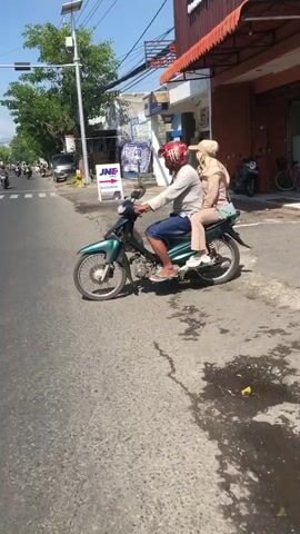 A man wearing a red helmet is riding a green motorcycle with a woman sitting behind him. The woman is wearing a beige headscarf and a beige outfit. They are on a paved road with businesses on the right side. A street light is visible on the left side of the road. The weather appears to be sunny.