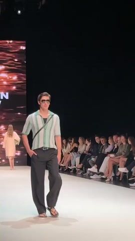 The video captures a male model walking down a runway. He is wearing a collared shirt with green and white stripes, black pants, and flip-flops. He also has on sunglasses and a small black bag slung across his body. The runway is white and well-lit, with audience members seated on either side. In the background, a large screen displays an abstract image, possibly of water. The overall atmosphere appears to be that of a fashion show.