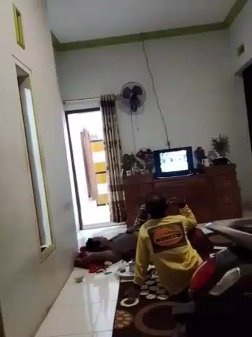The video shows an indoor scene, likely a living room, with two men visible. One man is lying on the floor, seemingly asleep or incapacitated, dressed in a dark shirt and pants. The second man is seated on the floor facing a television, wearing a yellow long-sleeved shirt with a graphic on the back. He appears to be looking at or interacting with the television, which is displaying a program. A fan is mounted on the wall above the television, and a wooden cabinet or entertainment center is positioned beneath it, holding various items, including plants and what appear to be cups. A door with curtains is visible on the left side of the frame, and part of a motorcycle or scooter is in the foreground on the right. The room has white tiled floors and light-colored walls with a green border near the ceiling.