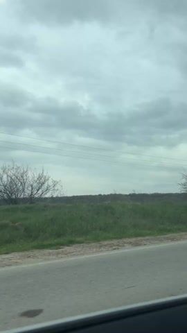 The video shows a car driving on a road on a cloudy, rainy day.  The view shifts from the side of the road, showing a grassy area with some trees, to the interior of the car, focusing on the windshield. Raindrops are visible on the windshield.  The road ahead is clear, with a white line dividing the lanes. A tall communication tower is visible in the distance, against a backdrop of green fields and low hills. The dashboard of the car is also visible, displaying time and other information.